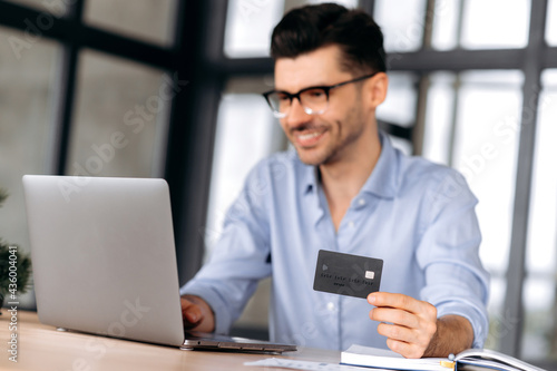 Blurred young modern Caucasian man wearing glasses, in casual clothes, sitting at table, using laptop and credit card to pay for online purchases or deliver goods home, smiling. Online shopping