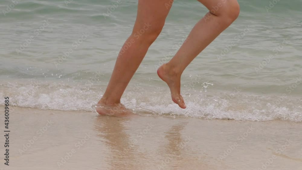 Woman run barefoot at swashing waves on sea beach, slow motion low half shot. Tracking camera shows only legs of happy and carefree tourist. Small waves move towards and break, water splash around Woman run barefoot at swashing waves on sea beach, slow motion low half shot. Tracking camera shows only legs of happy and carefree tourist. Small waves move towards and break, water splash around