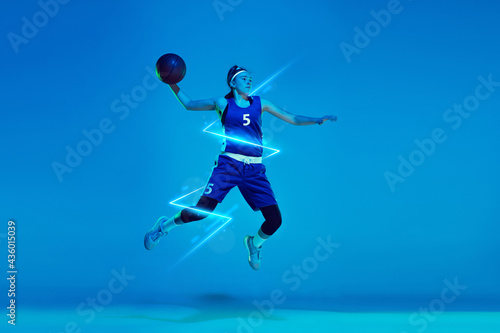 Young woman professional basketball player in jump isolated on blue studio background in neon light with geometric luminescent shapes, stripes © master1305
