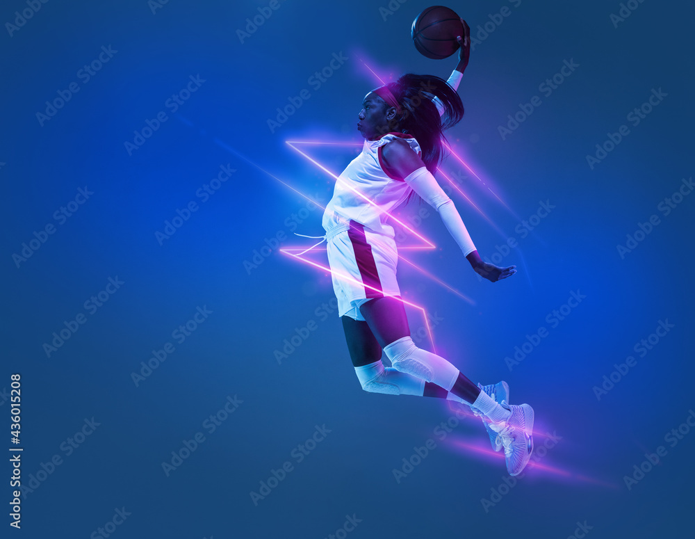 Young African woman basketball player in motion isolated on blue studio background in neon light with geometric luminescent shapes, stripes Young African woman basketball player in motion isolated on blue studio background in neon light with geometric luminescent shapes, stripes