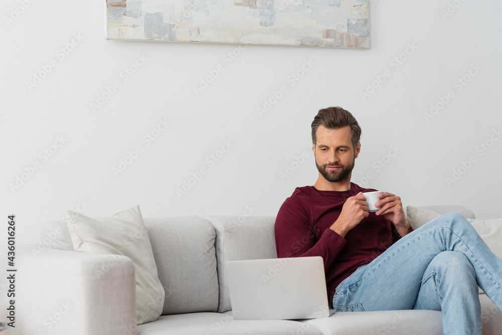 man with cup of coffee sitting on sofa near laptop