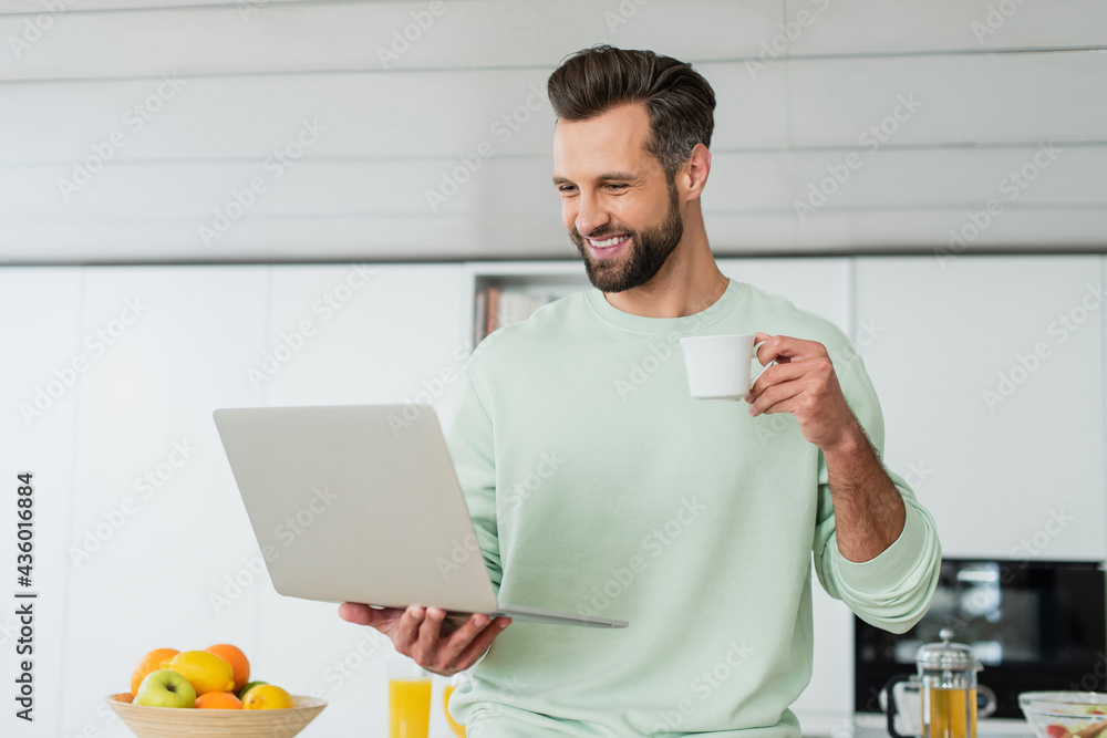 happy man looking at laptop while drinking coffee in kitchen happy man looking at laptop while drinking coffee in kitchen