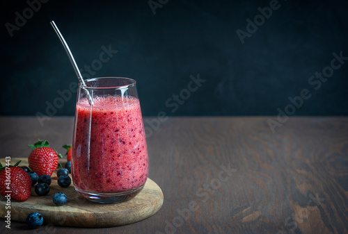 Refreshing organic smoothie made of juicy strawberries and blueberries served in drinking glass with metal reusable straw  on dark brown wooden table against black wall. Image with copy space photo