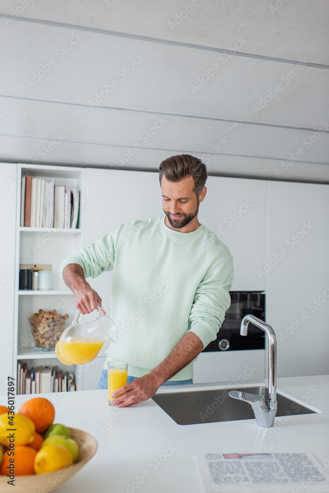 positive man pouring orange juice near fresh fruits on blurred foreground in kitchen positive man pouring orange juice near fresh fruits on blurred foreground in kitchen