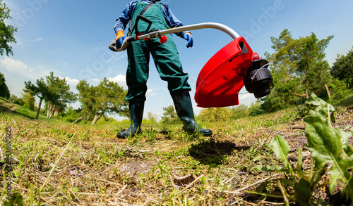 Photo of worker in green overalls and rubber boots using electric garden trimmer and cutting grass. photo