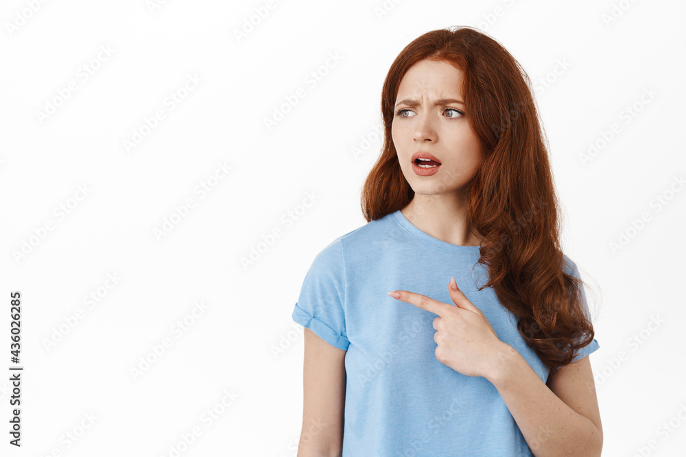 What is that. Confused young redhead woman frowning puzzled, pointing and looking left at something strange, stare puzzled at copy space for your logo or advertisement, white background What is that. Confused young redhead woman frowning puzzled, pointing and looking left at something strange, stare puzzled at copy space for your logo or advertisement, white background