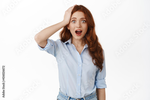 Image of surprised redhead woman remember something, pitching an idea, holding hand on head and staring in awe at camera, drop jaw, standing in blouse against white background © Cookie Studio