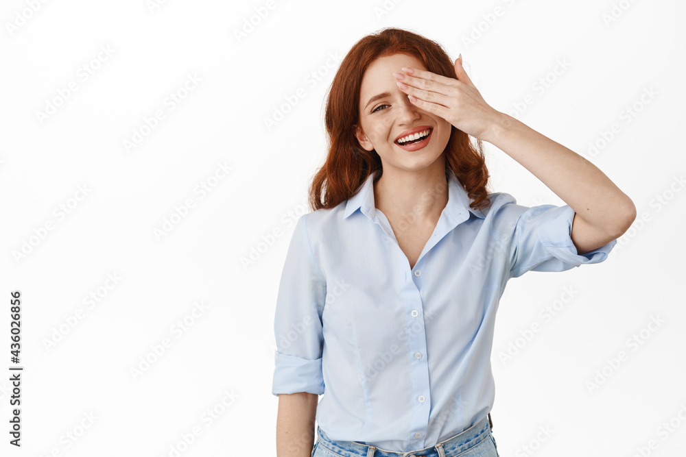 Carefree ginger girl cover one eye, smiling happy and sincere, feeling joy and happiness, standing in blouse against white background Carefree ginger girl cover one eye, smiling happy and sincere, feeling joy and happiness, standing in blouse against white background