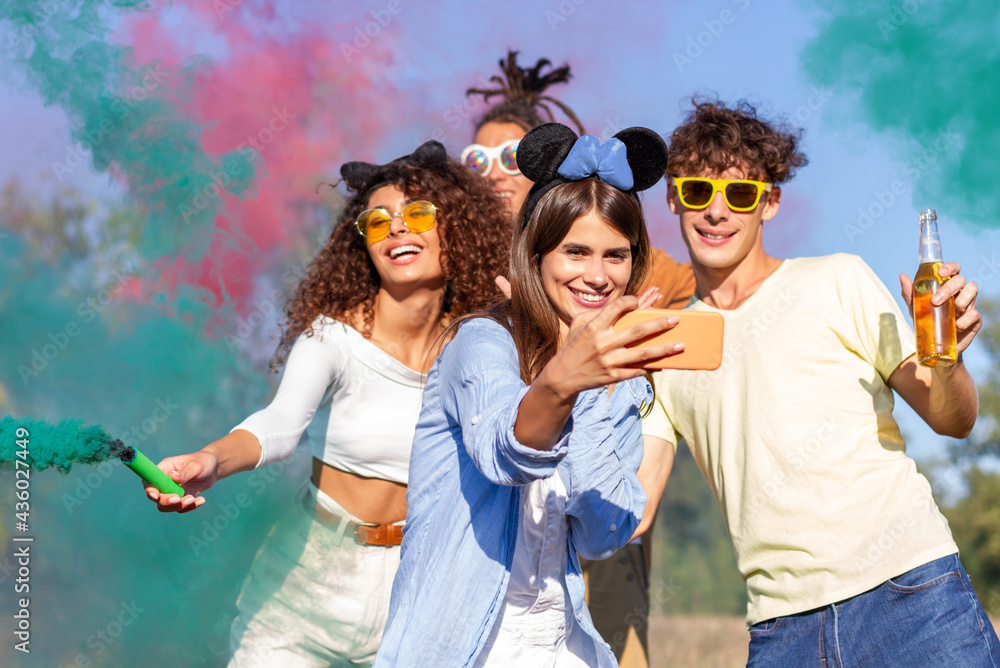 Beautiful young woman hold smartphone, doing selfie with happy friends,  having fun in the park with multicolored smoke bombs smoke fog. Young  students celebrating spring break together. Holi festival. Photos | Adobe