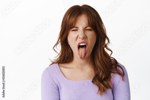 Portrait of disgusted young woman, grimacing from dislike, showing tongue disappointed with bad taste, awful smell, standing agaisnt white background