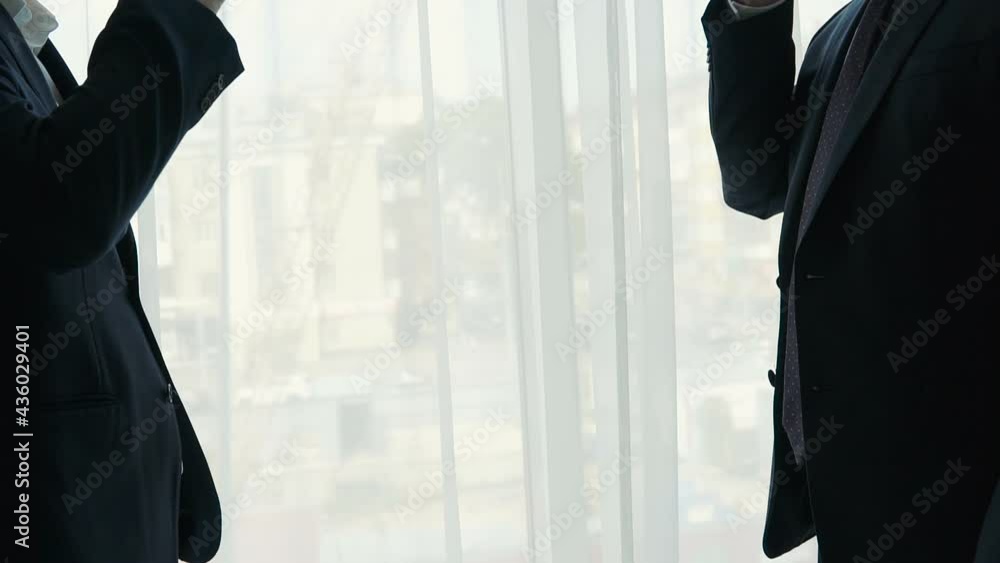 Silhouette of two men in business suits clinking cognac glasses. Toasting against window. Detail of businessmen hands raising toast after deal. Silhouette of two men in business suits clinking cognac glasses. Toasting against window. Detail of businessmen hands raising toast after deal.