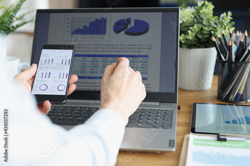 Employee holds smartphone with commercial business charts in his hand and compares them with image on laptop monitor