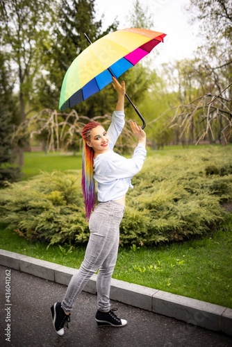 A cute girl with multi-colored braids and bright makeup in a bluish shirt posing with a rainbow umbrella against the background of a blooming spring park