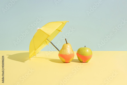 Summer fruit concept. Wet apple and a pear in monokini next to an umbrella isolated on a blue and yellow background. photo