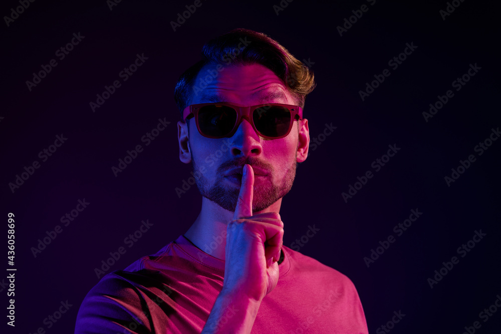 Portrait of attractive mysterious guy showing shh sign keep silence isolated over dark neon violet color background Portrait of attractive mysterious guy showing shh sign keep silence isolated over dark neon violet color background