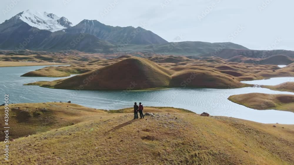 Aerial shot of a beautiful valley with surrounding mountains, two people on top of hill. River crossed green and brown valley during sunny day.Tagikistan