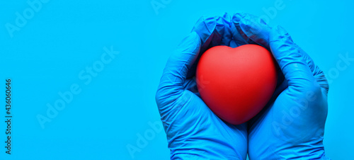 Both hands were wearing gloves and holding a red heart. Concept world heart foundation organizes world heart day, copy space on left for design or text, close-up, blurred blue background