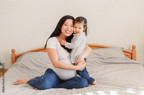 Asian Chinese pregnant woman with toddler girl hugging on bed at home. Girl daughter kid playing with mom. Mother and baby daughter expecting waiting for a new family member. Ethnic diversity