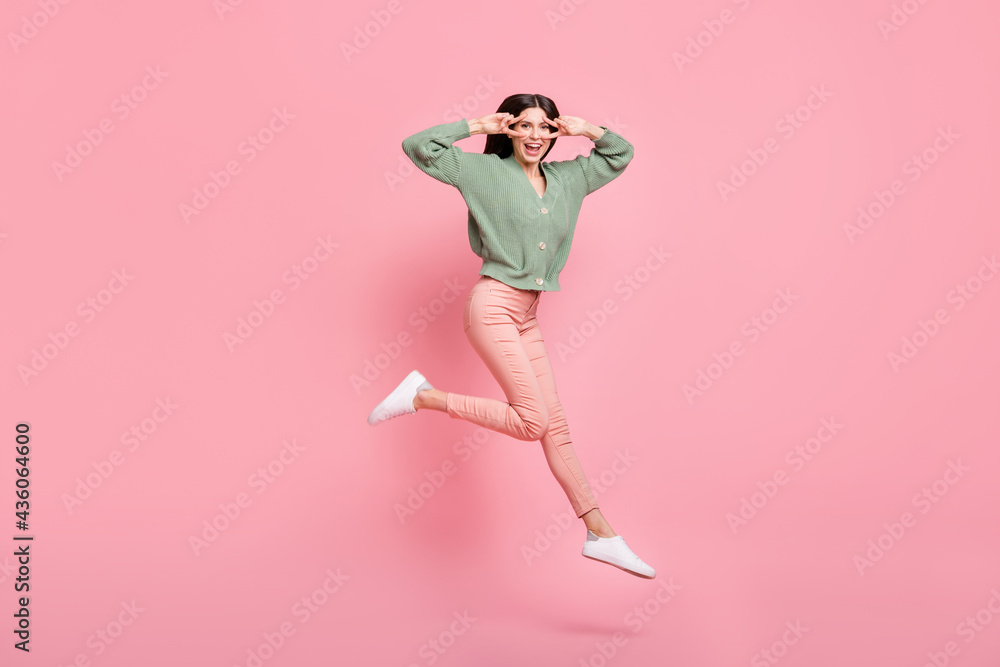Full length body size view of lovely funky cheerful girl jumping showing v-sign near eye having fun isolated over pink pastel color background Full length body size view of lovely funky cheerful girl jumping showing v-sign near eye having fun isolated over pink pastel color background