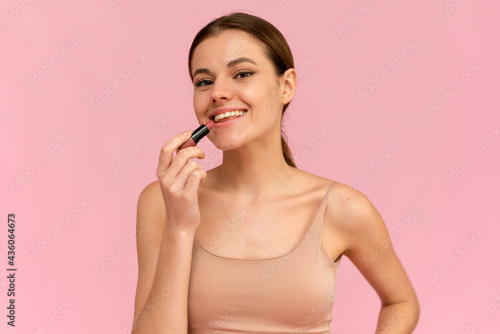 A young woman applies nude lipstick on her lips on a pink background. Cosmetics concept A young woman applies nude lipstick on her lips on a pink background. Cosmetics concept