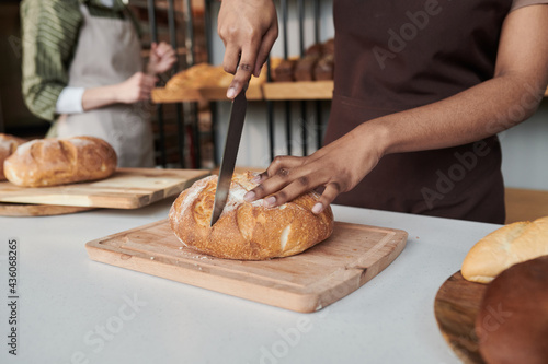 Close-up of worker in apron cutting fresh bread on cutting board at the table