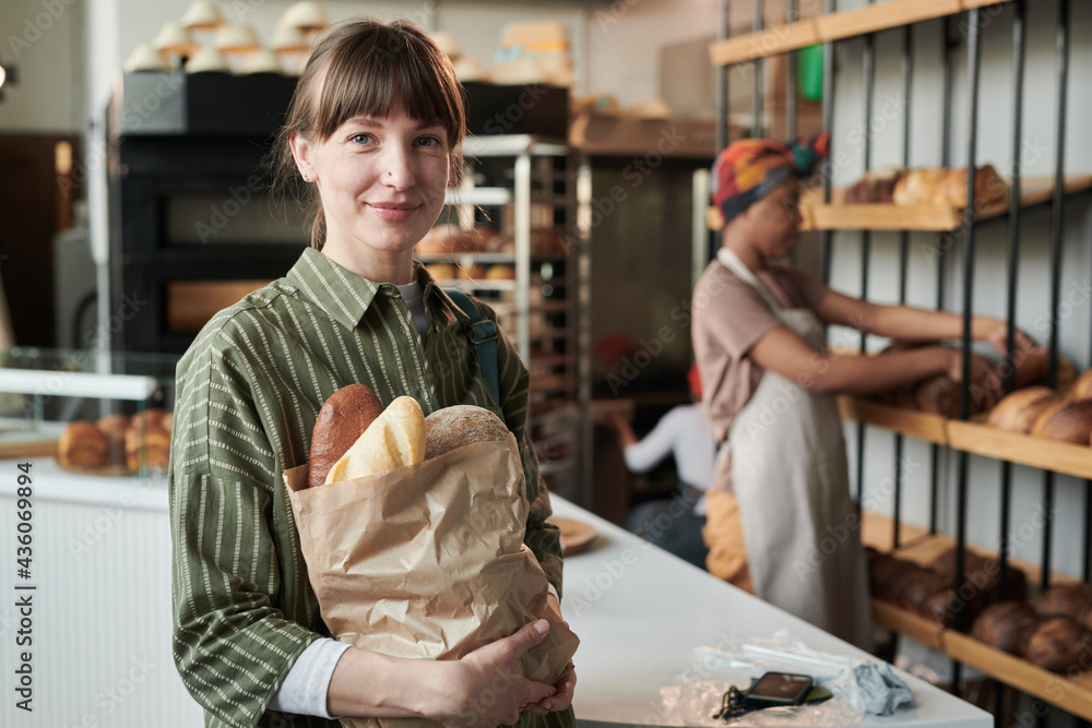 Portrait of young woman looking at camera and holding purchases she buying fresh bread in bakery shop