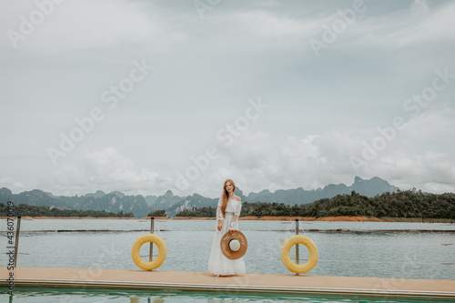 Happy Travel Girl with Hat Fun on Wooden Pier with Lake, Rainforest Jungle and Mountains on Background. Female Tourist Enjoy Vacation Cheow Lan Lake in Khao Sok National Park in Thailand. Trend Edit