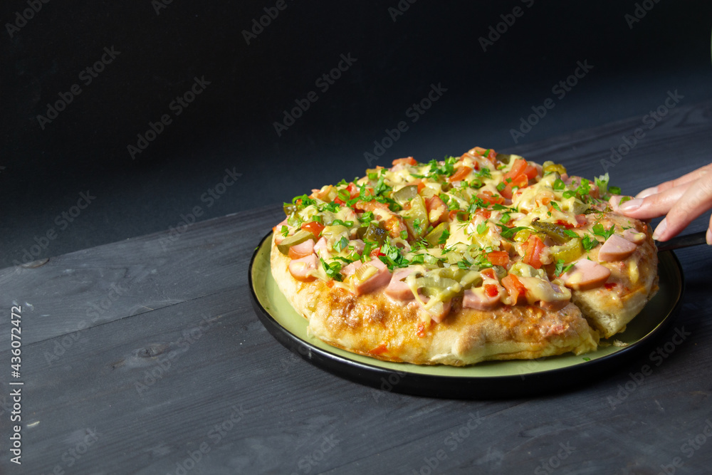 woman hamd holding slice of pizza with stretching cheese, sausage, marinated cucumbers, tomatoes and parsley on black background