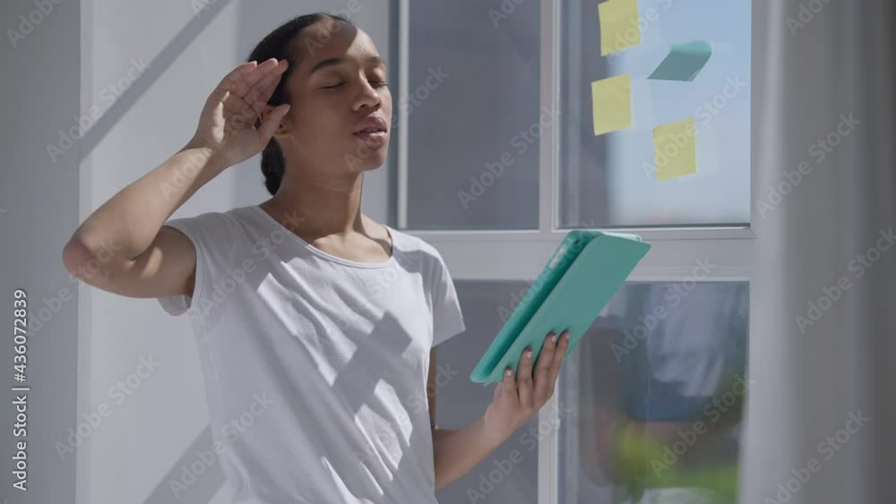 Sad fatigued worried African American woman wiping forehead looking at tablet screen standing in sunbeam at window. Portrait of tired overworked nervous lady waiting for e-mail in home office Sad fatigued worried African American woman wiping forehead looking at tablet screen standing in sunbeam at window. Portrait of tired overworked nervous lady waiting for e-mail in home office