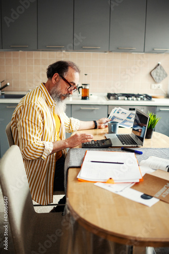 Side view of senior businessman in glasses calculating finances sitting at table in kitchen at home, open laptop, notepads and documents nearby