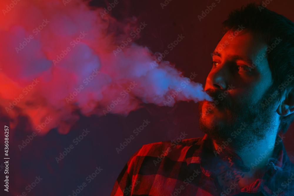 Tattooed man with a beard vaping and steaming his electronic cigarette. studio portrait with colored lights. alternative therapy for smoking cessation. Tattooed man with a beard vaping and steaming his electronic cigarette. studio portrait with colored lights. alternative therapy for smoking cessation.