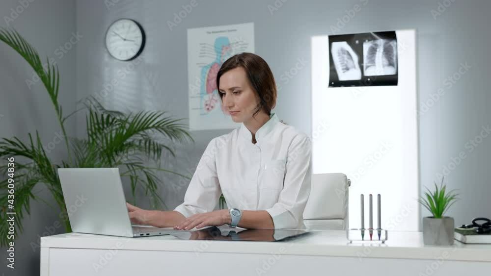 Female Doctor Sitting At The Table And Using Laptop. Young Woman In White Coat Sitting In The Office. Female Doctor Sitting At The Table And Using Laptop. Young Woman In White Coat Sitting In The Office.