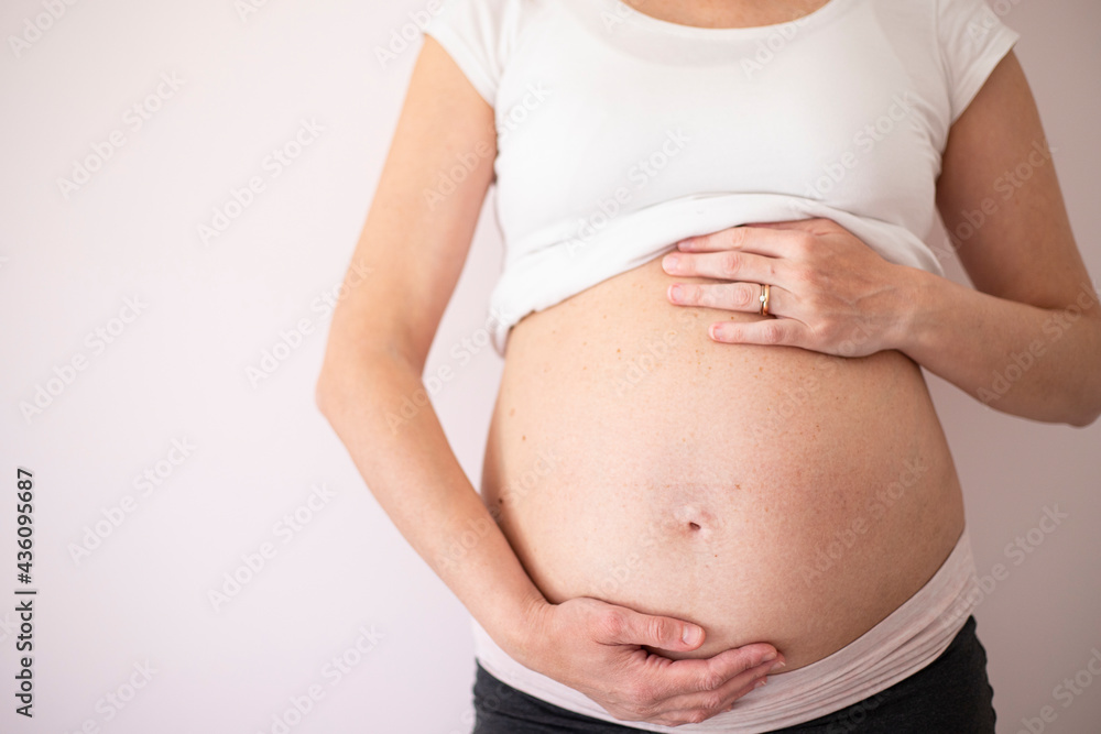 Pregnant woman wearing a white t-shirt with hands on belly on blank background. Central view. Background with copy space. Pregnant woman wearing a white t-shirt with hands on belly on blank background. Central view. Background with copy space.