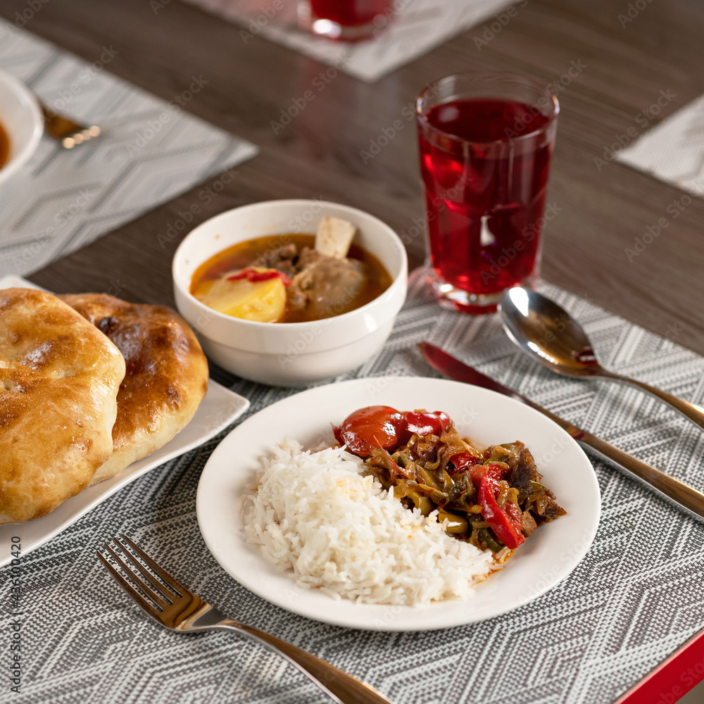 Set lunch from rice with vegetables, soup with meat broth, samsa and red compote on blurred background. Beautifully served dishes with delicious hearty food on wooden table with cutlery on it. Set lunch from rice with vegetables, soup with meat broth, samsa and red compote on blurred background. Beautifully served dishes with delicious hearty food on wooden table with cutlery on it.