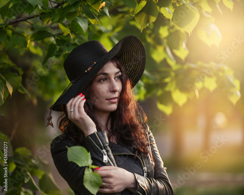 Close up portrait of Beautiful girl in dark clothet and black hat standing near green leaves on sunset. Art work of romantic woman. photo