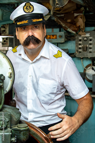 A sailor officer - old salt in the uniform is steering the ship with a rudder. Captain standing in the wheelhouse of ship and looking to camera.