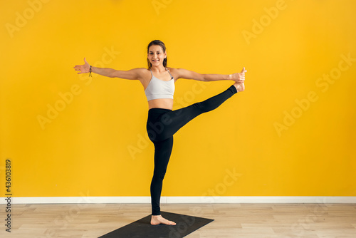 Young woman standing in yoga asana utthita hasta Padangusthasana, extended hand to big toe yoga pose , studio shot, yellow background