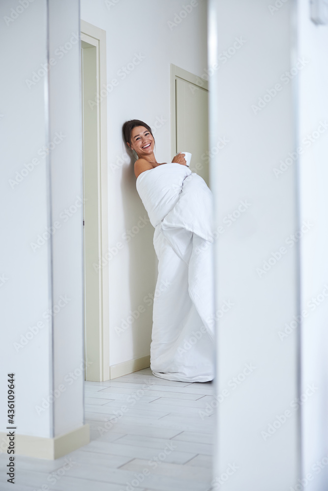 Pretty Caucasian lady covered in quilt while having fun in modern corridor in her flat