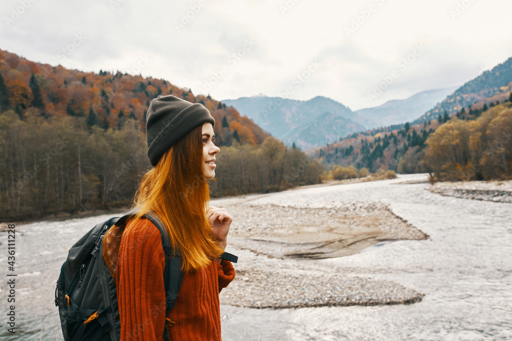 red-haired woman in a sweater with a backpack and in a hat near a mountain river in nature