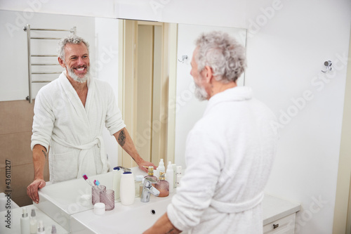 Happy smiling adult male looking to the reflection in the mirror in bathroom