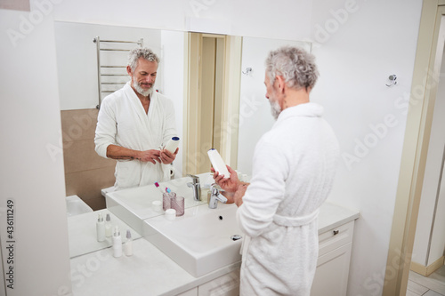 Handsome bearded male looking to the bottle with cosmetic in front of the mirror in bathroom