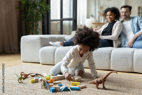 Small African American little girl child play with toy animals in living room, happy multiethnic mom and dad relax on sofa. Loving multiracial young family with daughter rest at home. Rental concept.