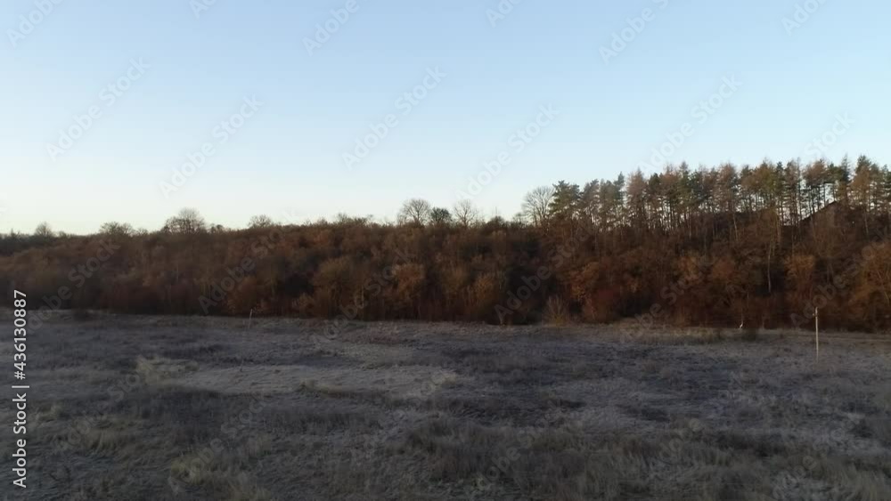 Early morning, the sun is rising over the village and small forest on the hill. Over the field covered with fog and the frost lies on the grass. The weather is fine and the sky is blue.