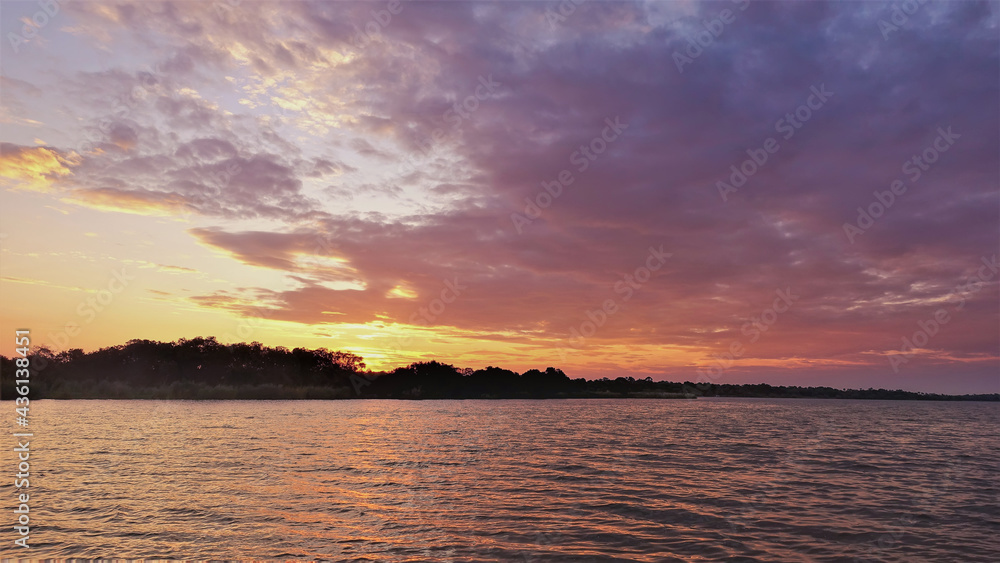 A flaming sunset over the Zambezi River. Clouds of purple and scarlet hues. The horizon is highlighted in gold. Reflections on the surface of the water. Zimbabwe