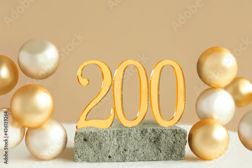 Number 200 on a concrete podium and volumetric golden balls on a beige background. Copy space..