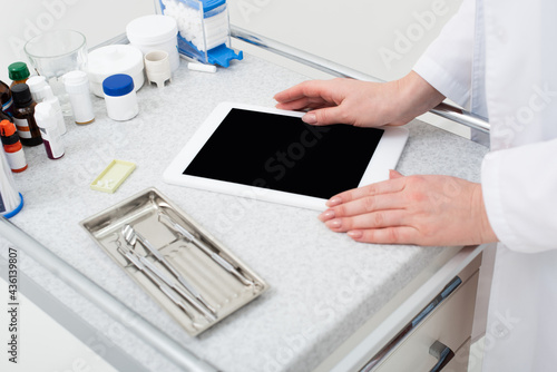 cropped view of female dentist hands using tablet with blank screen near metal dental tools in tray.