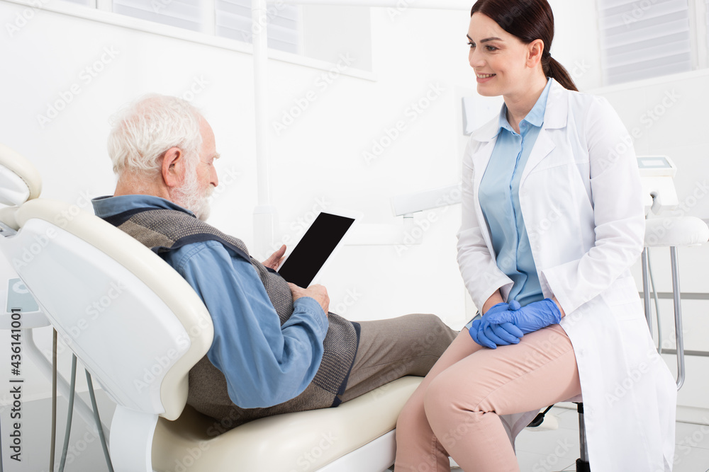 Obraz premium senior patient lying in dental chair and holding tablet with blank screen in hands near doctor in clinic.