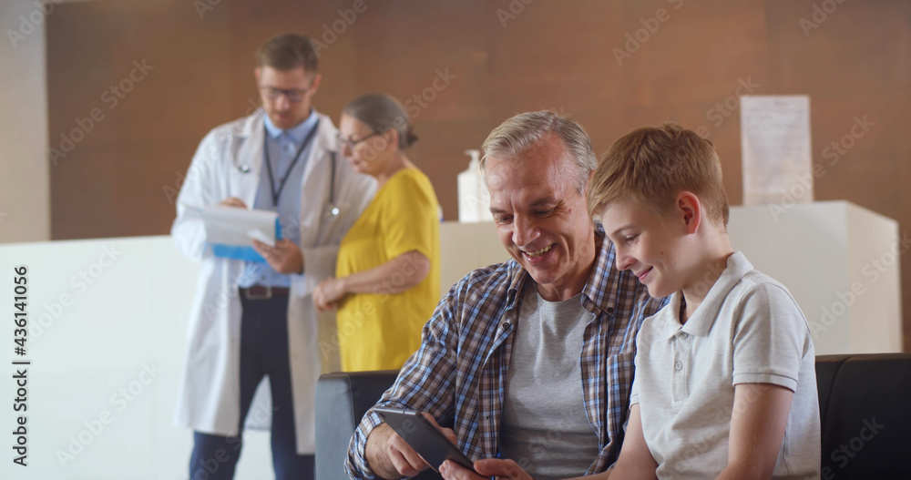 Obraz premium Father and son using digital tablet sitting in hospital waiting room