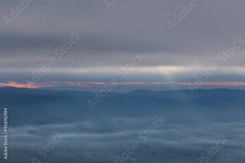 View from above of sunrays coming down on mist between hills and mountains in Umbria valley, Italy