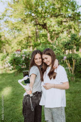 Smiling pretty young women influencer bloggers are filming or recording video with their mobile phone on a stabilizer, in a sunny green park outside. Blogging concept. Soft selective focus.